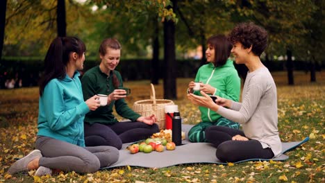 Happy-girls-are-having-picnic-in-park-sitting-on-yoga-mats-and-eating-after-outdoor-practice-in-autumn,-girls-are-talking-and-laughing.-Communication-and-food-concept.