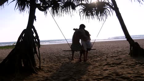 Young-romantic-couple-sitting-together-on-swing-at-the-tropical-sea-beach-and-hugging.-Pair-in-love-relaxing-and-kissing-on-ocean-exotic-shore-during-summer-vacation.-Holiday-concept-Back-rear-view
