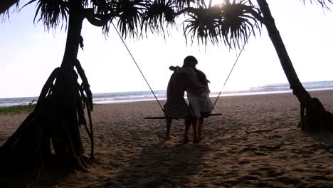 Joven-pareja-romántica-sentados-juntos-en-columpio-en-la-playa-del-mar-tropical-y-abrazos.-Par-en-amor-relajante-y-besos-en-la-exótica-costa-durante-las-vacaciones-de-verano.-Concepto-de-vacaciones-vista-posterior-posterior