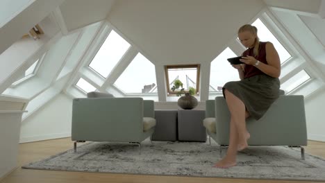 Vista-de-la-joven-mujer-rubia-sentada-en-el-lado-de-sillones-con-tablet-dentro-de-la-habitación-en-una-casa-cubo.-Rotterdam,-Países-Bajos