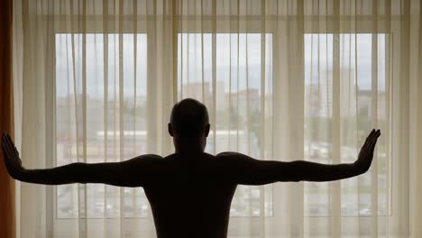 Man-infront-of-curtains-in-the-morning-raises-his-hands-and-stretch-oneself.-Adult-man-and-big-window-with-curtains.