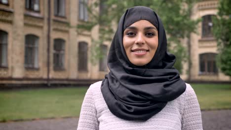 Young-beautiful-muslim-girl-in-hijab-is-standing-and-smiling-in-daytime,-in-bad-weather,-watching-at-camera,-building-on-background,-religiuos-concept