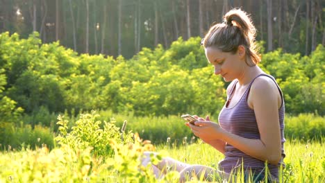 Chica-feliz-sentada-en-el-verde-césped-y-aplicaciones-smartphone-en-campo-escénico-en-el-fondo-de-la-puesta-del-sol