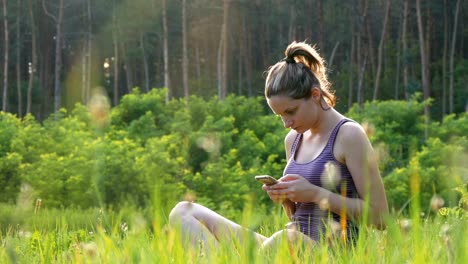 Happy-Young-Woman-Sitting-on-Green-Lawn-and-Uses-Smartphone-on-Scenic-Field-at-Sunset-Background