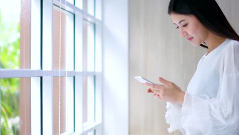 schöne-asiatische-Frau-genießen-Glück-Hand-Verwendung-Smartphone-in-der-Nähe-von-Fenster