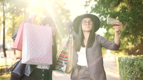 Chicas-despreocupadas-toman-selfie-con-smartphone-con-bolsas-en-la-calle-con-ropa-elegante.-Tecnología-moderna,-el-concepto-de-consumismo-y-la-diversión.