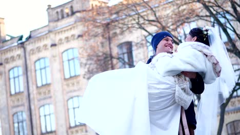 winter-wedding.-newlywed-couple-in-wedding-dresses.-groom-holds-bride-in-his-arms,-spinning.-they-are-happy,-smiling-at-each-other.-background-of-ancient-architecture,-snow-covered-park