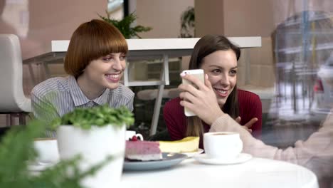 Three-young-caucasian-girls-are-sitting-in-a-cafe,-laughing,-smiling,-friends,-company,-gossips,-dialogue,-discussion.-Girlfriends-in-the-cafe-concept.-Using-a-smartphone,-watch-a-funny-video,-a-photo-60-fps