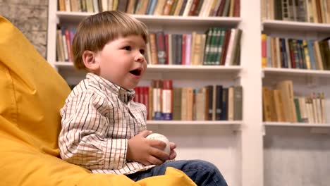 Little-boy-sitting-on-chair-and-catching-and-throwing-ball,-bookshelves-background