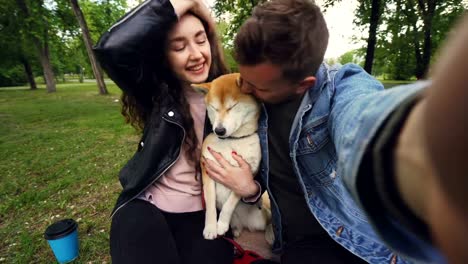 Point-of-view-shot-of-two-happy-people-and-puppy-taking-selfie-in-the-park,-kissing-and-loving-expressing-love-and-care.-Beautiful-green-lawn-and-trees-are-in-background.