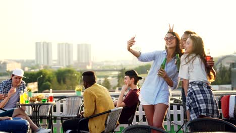 Mujeres-jóvenes-juguetones-están-tomando-selfie-con-smartphone-haciendo-muecas,-mostrando-la-lengua-y-orejas-de-conejo-que-se-divierten.-Chicas-vistiendo-ropa-de-moda-y-sosteniendo-botellas.