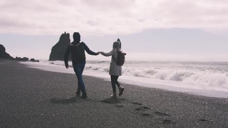 Pareja-joven-corriendo-por-la-playa-de-arena-negra-en-Islandia-y-divertirse,-cámara-lenta