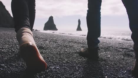 Close-up-shot-of-young-couple-holding-hands-walking-down-black-sand-beach-in-Iceland,-slow-motion