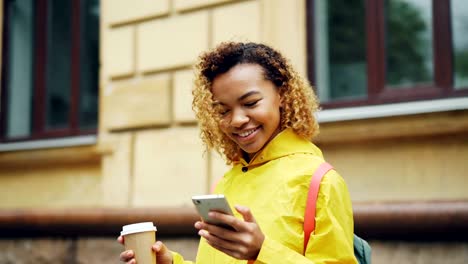 Pretty-African-American-girl-is-looking-at-smartphone-screen-and-touching-it-with-smile-standing-outdoors-wearing-bright-clothes-and-holding-coffee.-People-and-technology-concept.