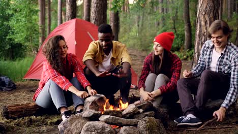 Young-men-and-women-travelers-are-sitting-around-fire,-telling-stories-and-laughing,-handsome-guy-is-throwing-firewood-in-flame.-Tent-and-backpacks-are-visible.