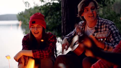 Multiracial-group-of-tourists-is-having-fun-in-forest-playing-the-guitar-and-singing-songs-around-campfire-in-wood-near-lake.-People-are-wearing-casual-clothing.