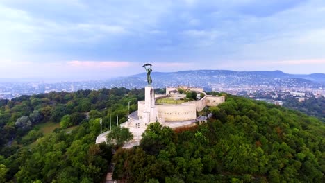 Herumfliegen-der-Statue-of-Liberty-mit-der-Skyline-von-Budapest-im-Hintergrund.-Budapest,-Ungarn---4K-Luftaufnahmen-bei-Sonnenaufgang