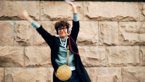 Mujer-joven-feliz-melómano-es-cantando-y-bailando-escuchando-música-en-auriculares-y-sostiene-el-teléfono-inteligente-al-aire-libre-en-la-calle.-Concepto-de-canciones-y-de-la-juventud.