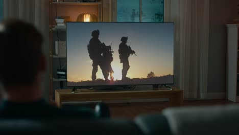 Man-Watches-Epic-War-Movie-on-TV-while-Sitting-on-a-Couch-at-Home-in-the-Evening.-Cozy-Living-Room-with-Warm-Lights.-Over-the-Shoulder-Shot.