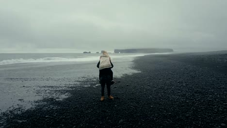 Aerial-view-of-the-young-couple-walking-on-the-black-volcanic-beach-in-Iceland.-Woman-in-lopapeysa-jumps-on-the-man-arms