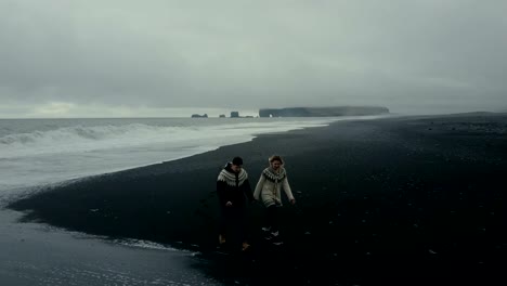 Aerial-view-of-young-couple-walking-on-the-shore-of-the-sea-in-Iceland.-Man-and-woman-look-to-the-wave-at-black-beach