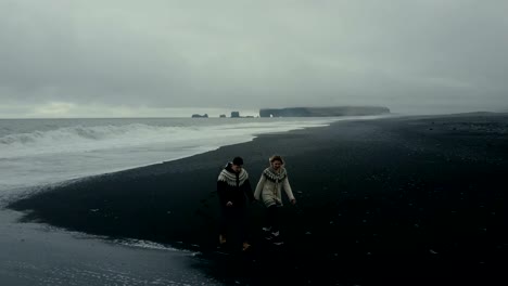 Vista-aérea-de-joven-pareja-caminando-en-la-orilla-del-mar-en-Islandia.-Hombre-y-la-mujer-mira-a-la-ola-en-playa-negra