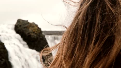 Portrait-of-young-beautiful-sad-woman-standing-near-the-waterfall-in-Iceland-and-looking-around.-Hair-waving-on-wind