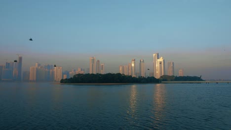 Locked-shot.-Sea-birds-fly-above-green-island-at-bay.-Skyscrapers-of-downtown-at-horizon.-Blue-sky-and-blue-water-lockdown-scene.