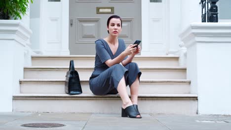 Elegante-mujer-sentada-sobre-las-medidas-del-edificio-mediante-teléfono-móvil