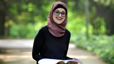 Portrait-of-a-young-girl-wearing-glasses-in-a-hijab-reading-a-book-in-nature,-laughing,-in-a-park-in-the-background.-50-fps
