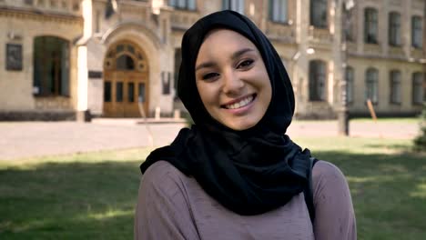 Young-beautiful-muslim-girl-in-hijab-is-standing-and-smiling-in-daytime-in-summer,-watching-at-camera,-building-on-background,-religiuos-concept