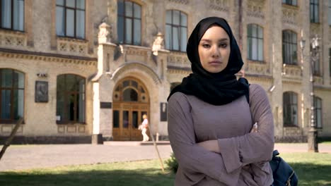 Young-serious-muslim-girl-is-standing-with-arms-crossed-in-daytime-in-summer,-watching-at-camera,-building-on-background,-religious-concept