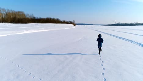 Aerial-footage-of-running-woman-in-winter-landscape-of-a-frozen-river-with-a-forest-and-a-view-of-the-city-with-factories.-Back-view.