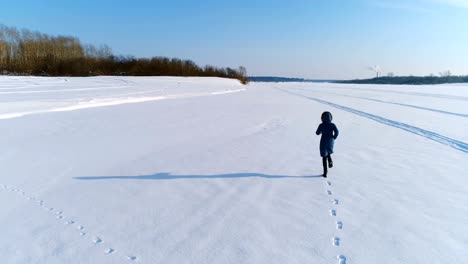 Luftaufnahmen-von-laufenden-Frau-in-Winterlandschaft-eines-gefrorenen-Flusses-mit-einem-Wald-und-einem-Blick-auf-die-Stadt-mit-Fabriken.-Hinten-Sie-Ansicht-von.