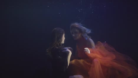 Couple-lesbian-woman-in-chiffon-dress-swimming-together-underwater-in-dark-pool