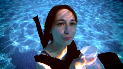 A-charming-young-girl-is-sitting-underwater-at-the-bottom-of-the-pool-in-a-beautiful-dress,-holding-a-white-Cup-and-saucer-in-her-hands,-looking-at-the-camera-and-smiling.-Vintage-processing.