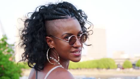 Trendy-young-black-woman-in-striped-camisole-and-sunglasses-walking-over-bridge,-looking-back-and-smiling-to-camera,-follow-shot