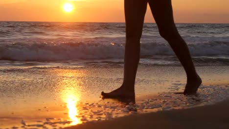 Legs-of-young-woman-going-along-ocean-beach-during-sunrise.-Female-feet-walking-barefoot-on-sea-shore-at-sunset.-Girl-stepping-in-shallow-water-at-shoreline.-Summer-vacation-concept.-Close-up