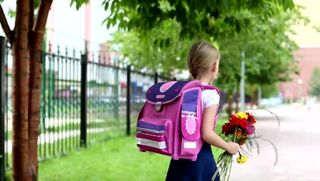 Schoolgirl-with-backpack,-flowers-bouquet-say-goodbye-and-go-away.-Girl-standing-near-school-with-bouquet-of-flowers-and-backpak.-Blured-unfocused-background.-Handheld-outdoors-shot-summer-day.