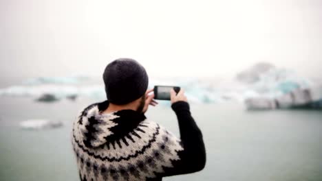 Young-stylish-man-standing-on-the-shore-in-Jokulsalon-ice-lagoon-in-Iceland-and-taking-photos-on-smartphone