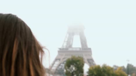 Young-beautiful-woman-walking-in-Paris,-France-and-taking-photos-of-Eiffel-tower-in-fog-on-smartphone-or-mobile-phone