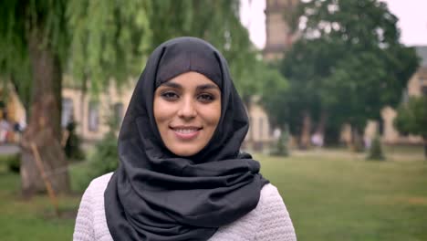 Young-beautiful-muslim-girl-in-hijab-is-standing-and-smiling-in-daytime,-in-bad-weather,-watching-at-camera,-building-on-background,-religiuos-concept,-blurred-background