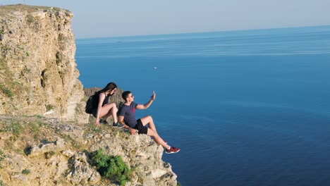 Junge-Backpacker-Frau-nähert-sich-Mann-sitzt-am-Rand-der-Felsen-paar-nehmen-selfie