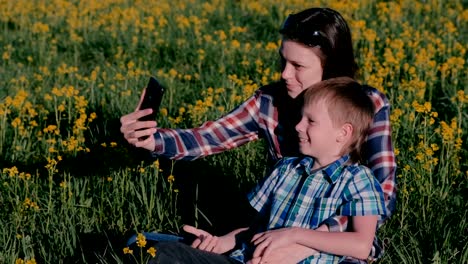 Mutter-und-Sohn-machen-Selfie-am-Telefon-sitzen-auf-dem-Rasen-unter-den-gelben-Blumen.