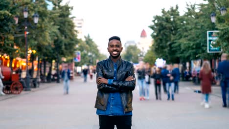 Zoom-in-time-lapse-of-happy-African-American-guy-wearing-jeans-and-leather-jacket-standing-alone-in-street-downtown,-smiling-and-looking-at-camera-with-crowd-moving-by.