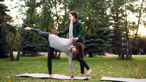 Instructora-de-yoga-amable-ayuda-a-estudiante-femenino-para-mantener-la-pose-de-media-luna-Ardha-Chandrasana-junto-a-ella,-hablando-y-mantenerla-a-mano-y-la-pierna.