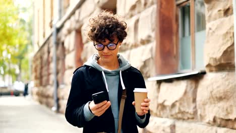 Beautiful-girl-with-dark-curly-hair-is-using-smartphone-texting-friends-and-holding-coffee-walking-in-city-alone.-Modern-technology,-communication-and-drinks-concept.