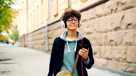 Fröhliche-junge-Dame-ist-Musik-über-Kopfhörer-hören-und-Smartphone-auf-Straße-Herbsttag-alleine-halten.-Kultur-und-Technik-Jugendkonzept.