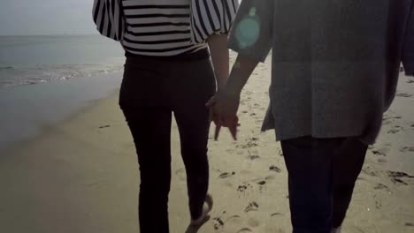 Two-women-holding-hands-together-while-walking-on-sandy-beach.