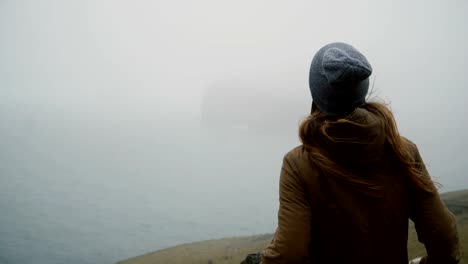 Back-view-of-young-woman-in-hat-standing-on-the-shore-of-the-sea-and-thinking-about-something,-exploring-the-Iceland